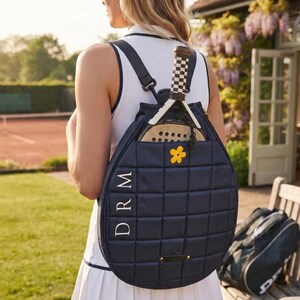May include: A navy blue quilted backpack with a vertical grid pattern, featuring the white letters DRM and a small yellow flower icon. A paddle with a black and white checkered handle is tucked into the top compartment of the bag.