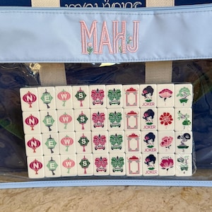 May include: A Mahjong set with a blue and white tote bag. The tiles are white with various symbols in green, pink, and red. The bag has the word "MAHJ" embroidered in pink. The set is visible through a clear plastic window.