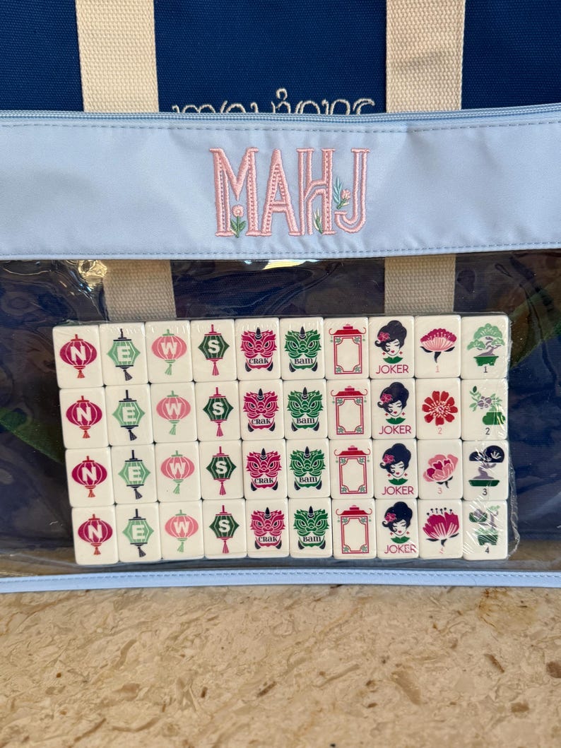 May include: A Mahjong set with a blue and white tote bag. The tiles are white with various symbols in green, pink, and red. The bag has the word "MAHJ" embroidered in pink. The set is visible through a clear plastic window.