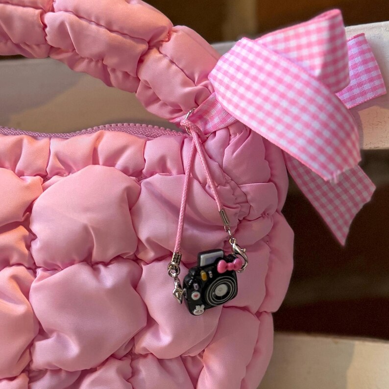 May include: A pink quilted handbag with a matching pink gingham bow. The bag features a small black camera charm with a pink bow. The bag's handle is visible at the top, and the zipper is partially visible.