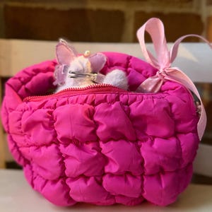 May include: A bright pink, quilted, zippered bag with a pink ribbon bow. Inside the bag is a small, plush, lavender-colored toy with a silver clasp. The bag is sitting on a white surface.