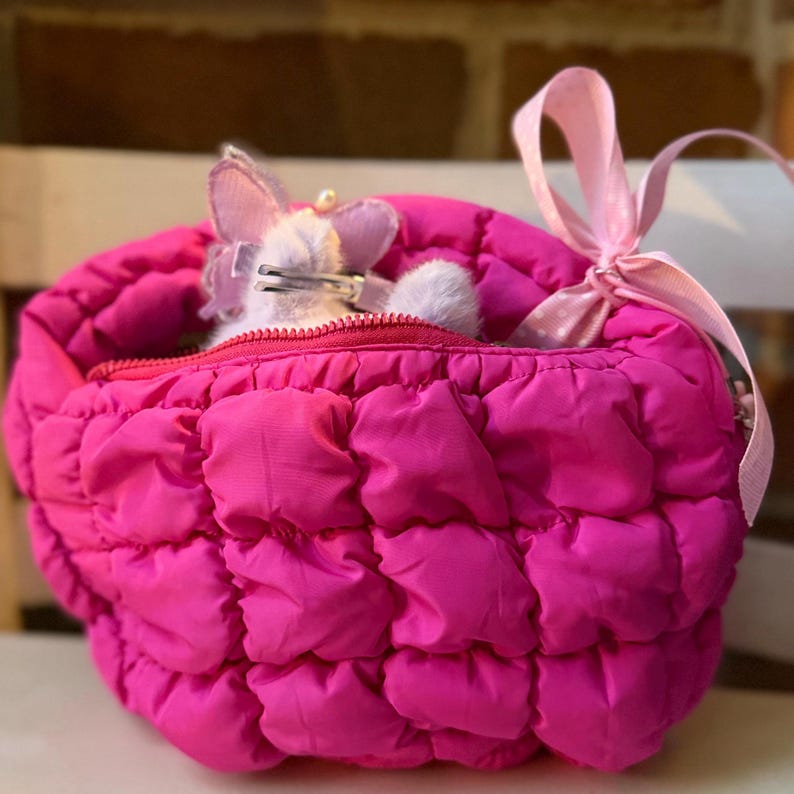 May include: A bright pink, quilted, zippered bag with a pink ribbon bow. Inside the bag is a small, plush, lavender-colored toy with a silver clasp. The bag is sitting on a white surface.