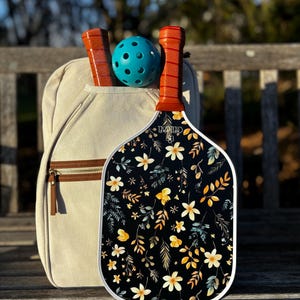 K&ouml;nnte beinhalten: Ein wei&szlig;-schwarzer Pickleball-Schl&auml;ger mit Blumenmuster. Der Schl&auml;ger hat einen braunen Griff und liegt auf einer Holzbank. Im Hintergrund befindet sich ein blauer Pickleball.