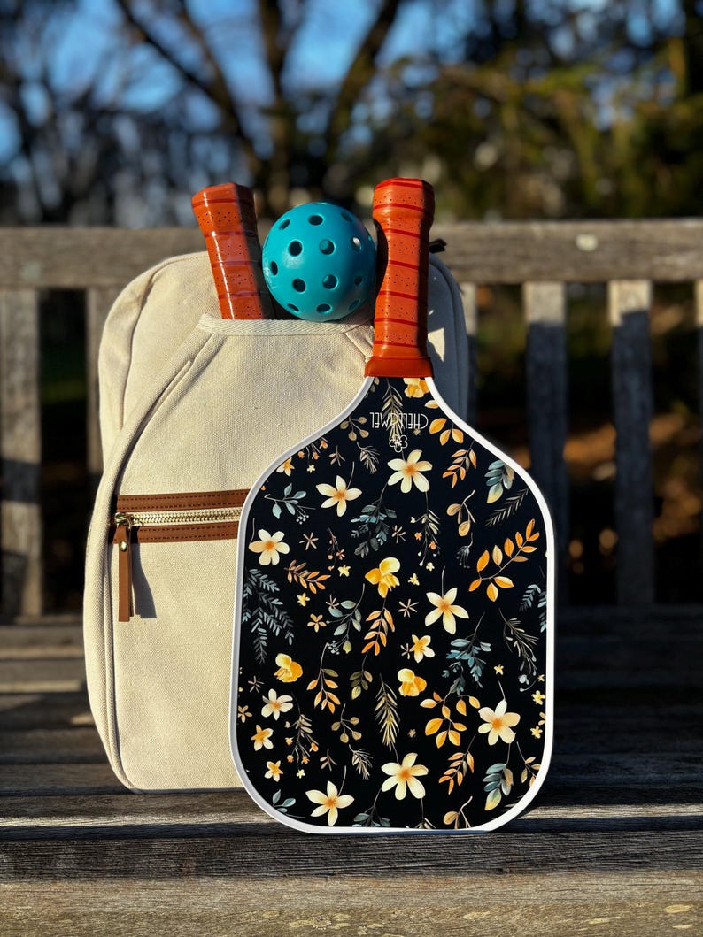 K&ouml;nnte beinhalten: Ein wei&szlig;-schwarzer Pickleball-Schl&auml;ger mit Blumenmuster. Der Schl&auml;ger hat einen braunen Griff und liegt auf einer Holzbank. Im Hintergrund befindet sich ein blauer Pickleball.