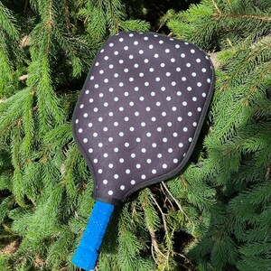 Pu&ograve; includere: Una racchetta da pickleball a pois neri e bianchi con un manico blu. La racchetta &egrave; appoggiata su un letto di aghi di pino verdi.