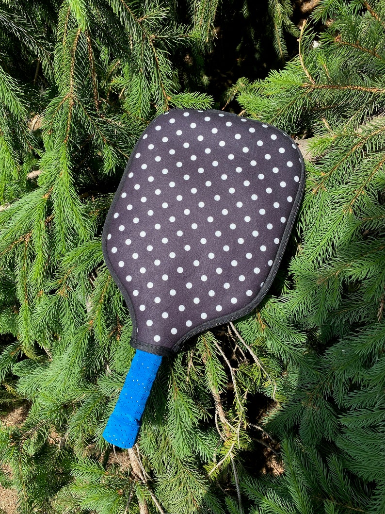 Pu&ograve; includere: Una racchetta da pickleball a pois neri e bianchi con un manico blu. La racchetta &egrave; appoggiata su un letto di aghi di pino verdi.