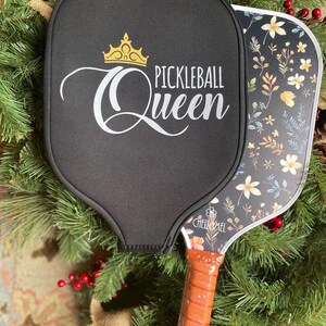 K&ouml;nnte beinhalten: Ein schwarzer Pickleball-Schl&auml;ger mit floralem Design und einer goldenen Krone mit dem Text "Pickleball Queen" auf der Schl&auml;gerfl&auml;che. Der Schl&auml;ger hat einen braunen Griff.