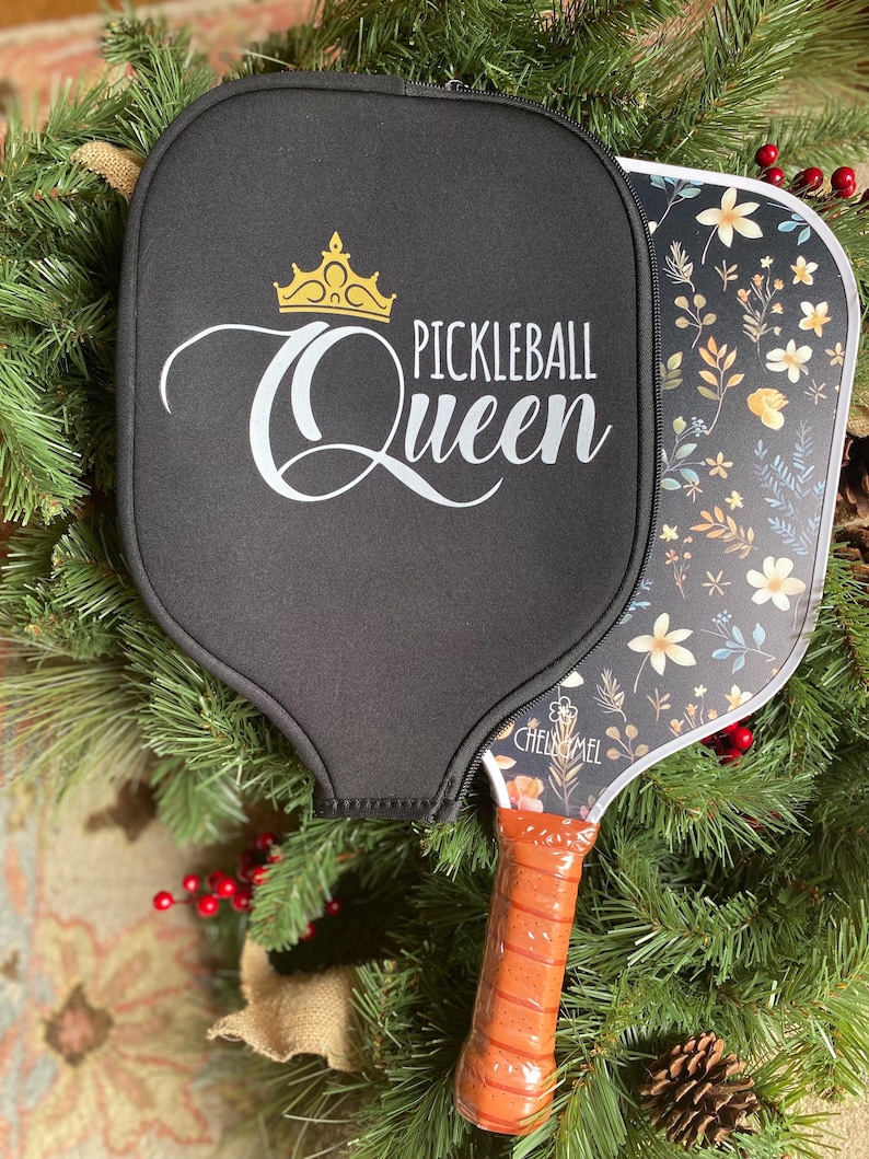 K&ouml;nnte beinhalten: Ein schwarzer Pickleball-Schl&auml;ger mit floralem Design und einer goldenen Krone mit dem Text "Pickleball Queen" auf der Schl&auml;gerfl&auml;che. Der Schl&auml;ger hat einen braunen Griff.