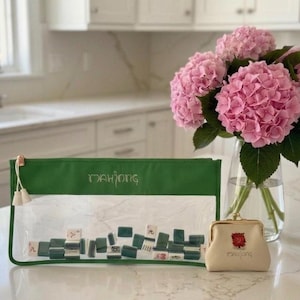 May include: A clear, rectangular Mahjong tile case with a green top and gold zipper, filled with tiles. A small, beige coin purse with a red floral design and the word "Mahjong" is next to the case. A vase of pink hydrangeas is in the background.
