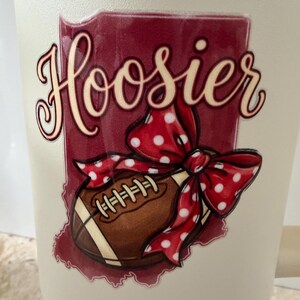 May include: A white tumbler with a maroon graphic featuring the word "Hoosier" in a script font. A brown football with white stripes and a red polka dot bow are also featured in the graphic.