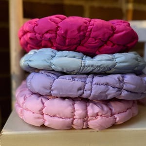 May include: A stack of four padded headbands in shades of pink, purple, and blue. The headbands are made of a quilted fabric and appear soft and comfortable. The headbands are stacked on top of each other, with the pink headband on top.