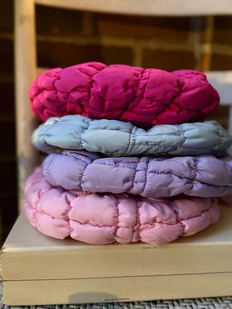 May include: A stack of four padded headbands in shades of pink, purple, and blue. The headbands are made of a quilted fabric and appear soft and comfortable. The headbands are stacked on top of each other, with the pink headband on top.