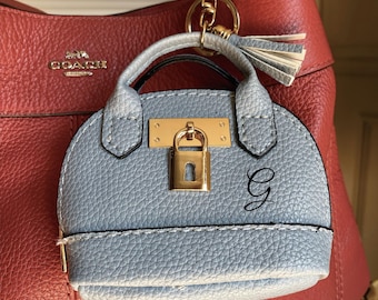 May include: A light blue purse charm with a gold-colored lock and handle, attached to a gold keychain. The charm has a small tassel and the letter "G" on the front. The charm is hanging on a red leather purse.