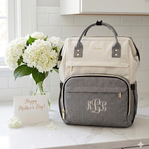 May include: A cream and gray diaper bag with gold zippers and a monogram. The bag has a top handle and a front pocket. A card that says "Happy Mother's Day!" is next to a vase of white flowers.