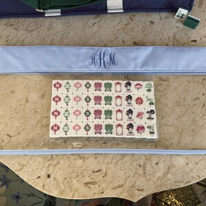 May include: A clear, rectangular Mahjong tile case with a light blue trim and a monogram. Inside the case are several Mahjong tiles with various symbols and characters. A blue tote bag is visible in the background.