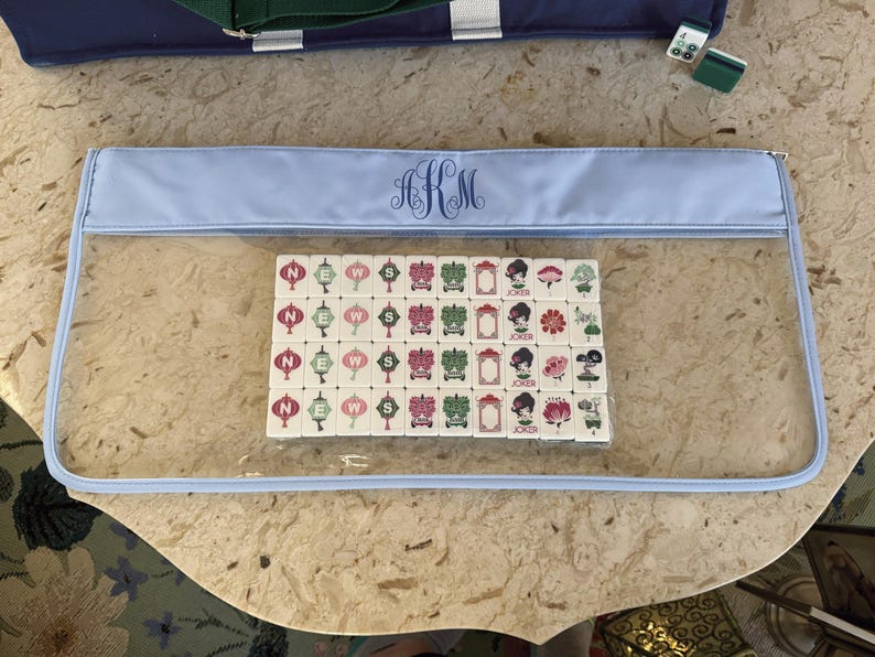 May include: A clear, rectangular Mahjong tile case with a light blue trim and a monogram. Inside the case are several Mahjong tiles with various symbols and characters. A blue tote bag is visible in the background.