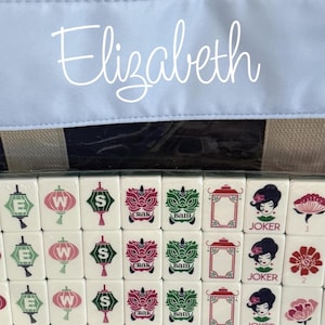 May include: A close-up of a Mahjong set with white tiles featuring various symbols and characters. The word "Elizabeth" is written in cursive on a light blue background. The set is partially visible.