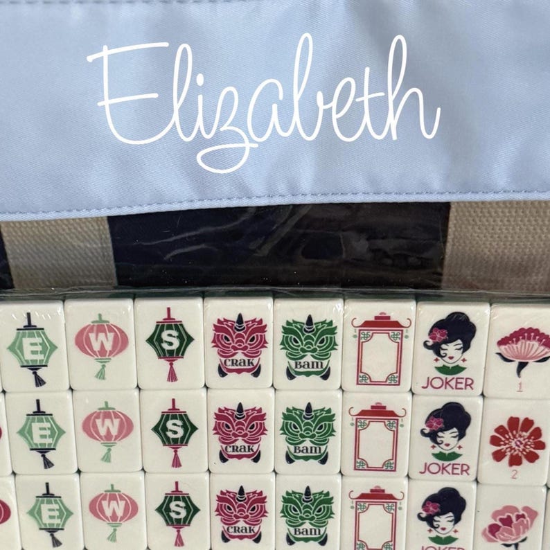 May include: A close-up of a Mahjong set with white tiles featuring various symbols and characters. The word "Elizabeth" is written in cursive on a light blue background. The set is partially visible.