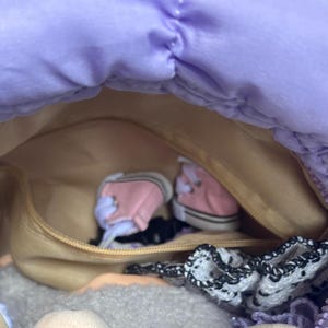 May include: A lavender quilted handbag with a zippered closure, revealing a tan interior. Inside are miniature pink sneakers, a black hair tie, and a white and black lace accessory. The bag is open, showcasing its contents.