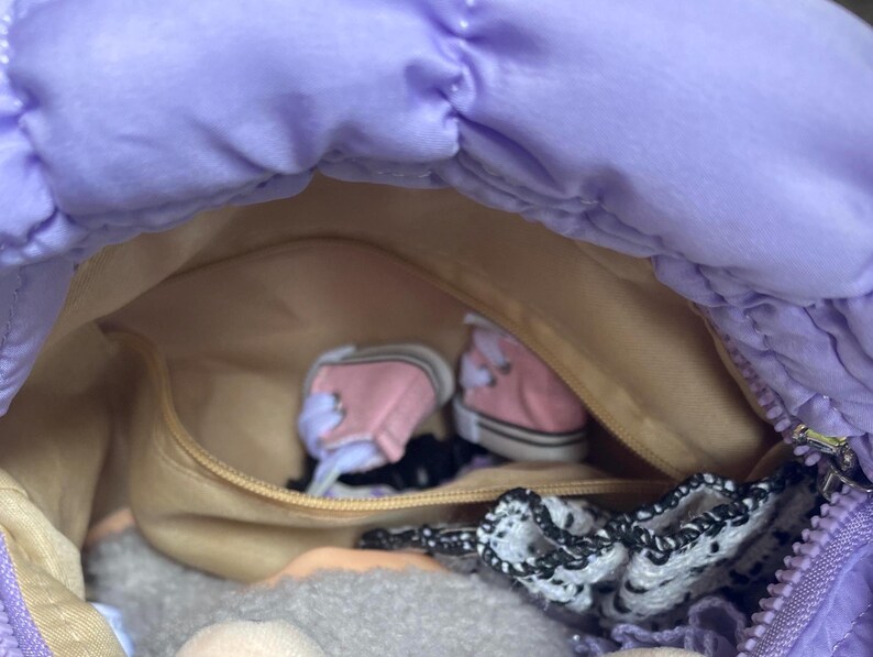 May include: A lavender quilted handbag with a zippered closure, revealing a tan interior. Inside are miniature pink sneakers, a black hair tie, and a white and black lace accessory. The bag is open, showcasing its contents.