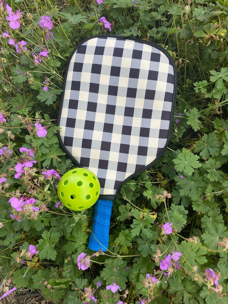 Pu&ograve; includere: Una racchetta da paddleball a scacchi bianco e nero con un manico blu e una pallina da paddleball verde.