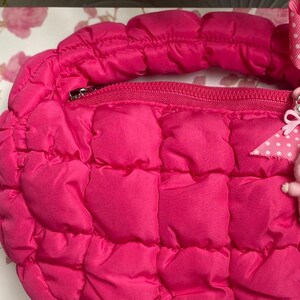May include: A quilted, hot pink handbag with a zipper closure and a matching handle. The bag has a puffy, padded texture and a small, decorative charm attached to the zipper.