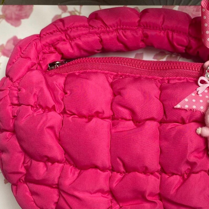 May include: A quilted, hot pink handbag with a zipper closure and a matching handle. The bag has a puffy, padded texture and a small, decorative charm attached to the zipper.