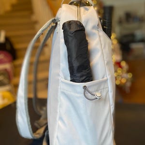 Op de afbeelding: Een lichtgrijze rugzak met een zwarte paraplu in de zijzak. De rugzak heeft een ritssluiting en grijze banden. Een tennisracket is zichtbaar in de tas.