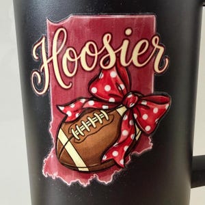 May include: Black coffee mug featuring a design with the word "Hoosier" in a script font, a football, and a red polka dot bow. The design is set against a maroon outline of the state of Indiana.