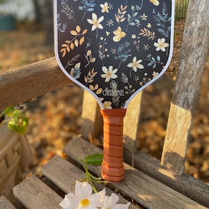 K&ouml;nnte beinhalten: Ein wei&szlig;-schwarzer Schl&auml;ger mit Blumenmuster. Der Schl&auml;ger hat einen braunen Griff und der Text "Chell&Mel" ist auf dem Schl&auml;ger aufgedruckt.