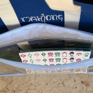 May include: A blue and white striped tote bag with the word "Mahjong" embroidered in white. Inside, a clear zippered pouch holds a set of Mahjong tiles with colorful designs.