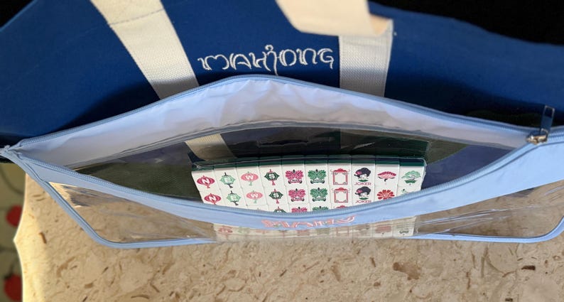 May include: A blue and white striped tote bag with the word "Mahjong" embroidered in white. Inside, a clear zippered pouch holds a set of Mahjong tiles with colorful designs.