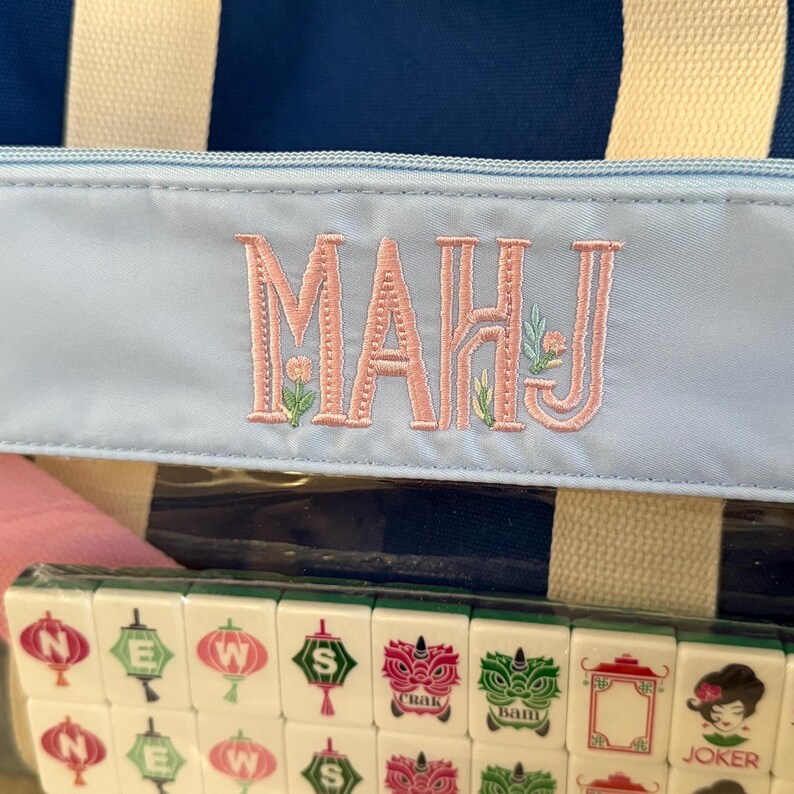 May include: A blue and white striped tote bag with a light blue zippered pocket. The pocket features the embroidered letters "MAHJ" in pink with floral accents. Below the bag is a set of mahjong tiles.