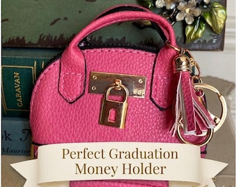 May include: A bright pink purse-shaped money holder with a gold-colored lock and handle. The purse has a textured surface and a tassel keychain. The text on the banner reads "Perfect Graduation Money Holder".