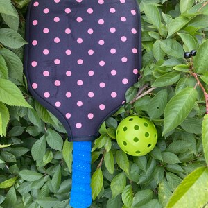 Pu&ograve; includere: Una racchetta da pickleball nera e rosa a pois con un manico blu e una palla da pickleball verde.