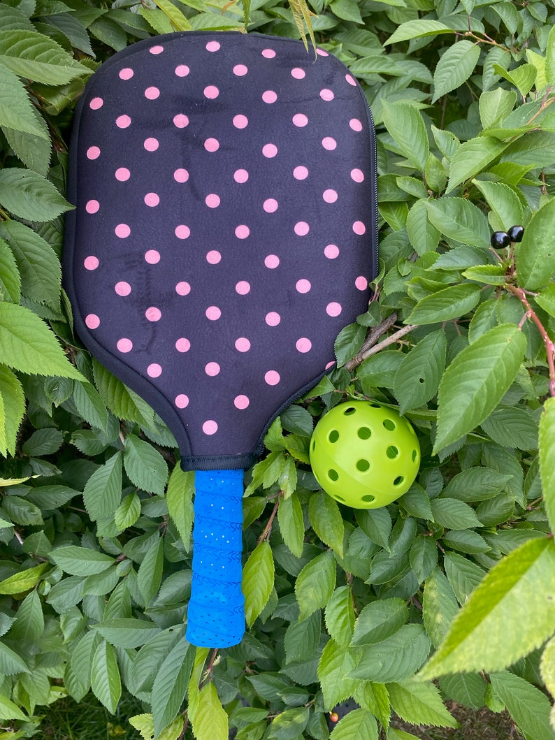 Pu&ograve; includere: Una racchetta da pickleball nera e rosa a pois con un manico blu e una palla da pickleball verde.