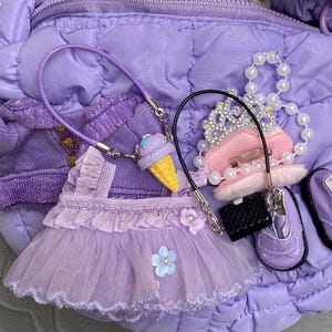 May include: A collection of miniature purple doll accessories, including a dress, jacket, shoes, purse, tiara, and a necklace with an ice cream cone charm. The items are arranged on a quilted purple surface.