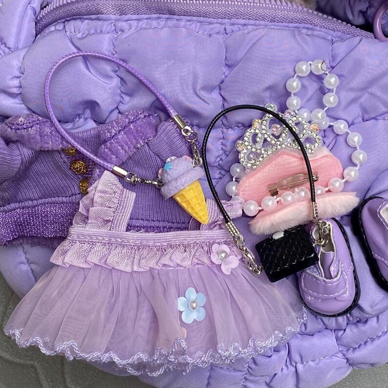 May include: A collection of miniature purple doll accessories, including a dress, jacket, shoes, purse, tiara, and a necklace with an ice cream cone charm. The items are arranged on a quilted purple surface.
