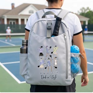 May include: A light gray backpack featuring a graphic print of figures playing pickleball and the text "Dink on Style." The bag includes a top handle, a vertical zippered pocket, a side water bottle sleeve, and a mesh side pocket holding two blue balls.