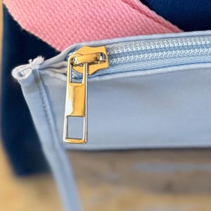May include: Close-up of a light blue bag with a gold zipper. A pink strap is visible, along with a navy blue background. The zipper is open, revealing the interior of the bag.