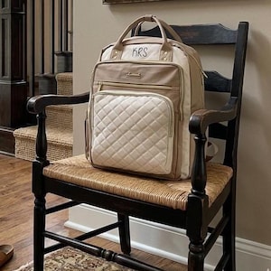 May include: A beige and cream-colored diaper bag with gold zippers and the initials "KRS" embroidered on the top. The bag has a quilted front pocket and is sitting on a dark wooden chair with a woven seat.