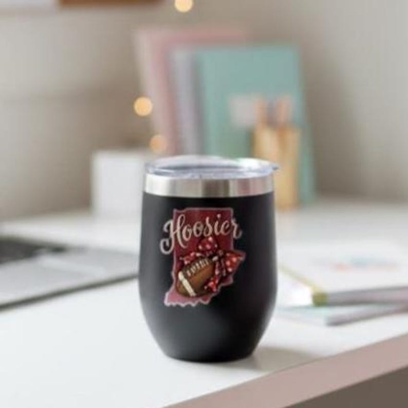 May include: A black insulated tumbler with a silver rim and a clear lid. The tumbler features a graphic of the state of Indiana with the word "Hoosier" and a football design. The tumbler is designed to keep beverages cold or hot.