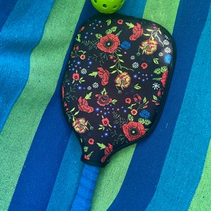 Pu&ograve; includere: Una racchetta da pickleball nera e blu con un motivo floreale sulla faccia della racchetta. La racchetta &egrave; appoggiata su un asciugamano a strisce blu e verdi con una palla da pickleball verde sopra l'asciugamano.