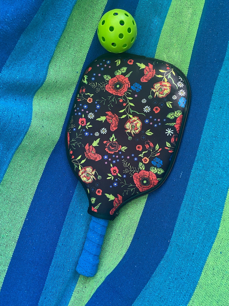 Pu&ograve; includere: Una racchetta da pickleball nera e blu con un motivo floreale sulla faccia della racchetta. La racchetta &egrave; appoggiata su un asciugamano a strisce blu e verdi con una palla da pickleball verde sopra l'asciugamano.