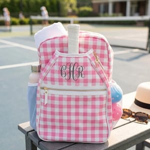 May include: A pink and white gingham backpack featuring a front pocket with a dark gray monogram of the letters CHR. The bag includes a white tennis racket handle protruding from the top, a side mesh pocket with two balls, and gold zippers.