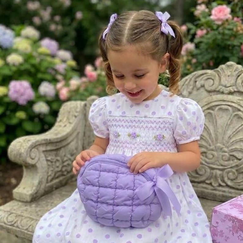 May include: A young girl wearing a white dress with purple polka dots and puff sleeves, holding a quilted lavender purse with a matching bow. The dress has a smocked bodice with floral embroidery. The girl has two purple bows in her hair.