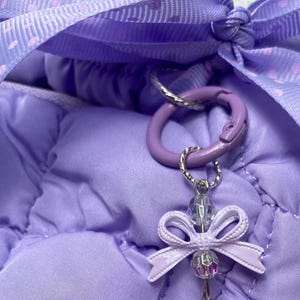 May include: Close-up of a lavender-colored bag with a matching ribbon and a decorative charm. The charm features a small bow and iridescent beads, attached to a purple carabiner clip. The bag has a quilted texture.