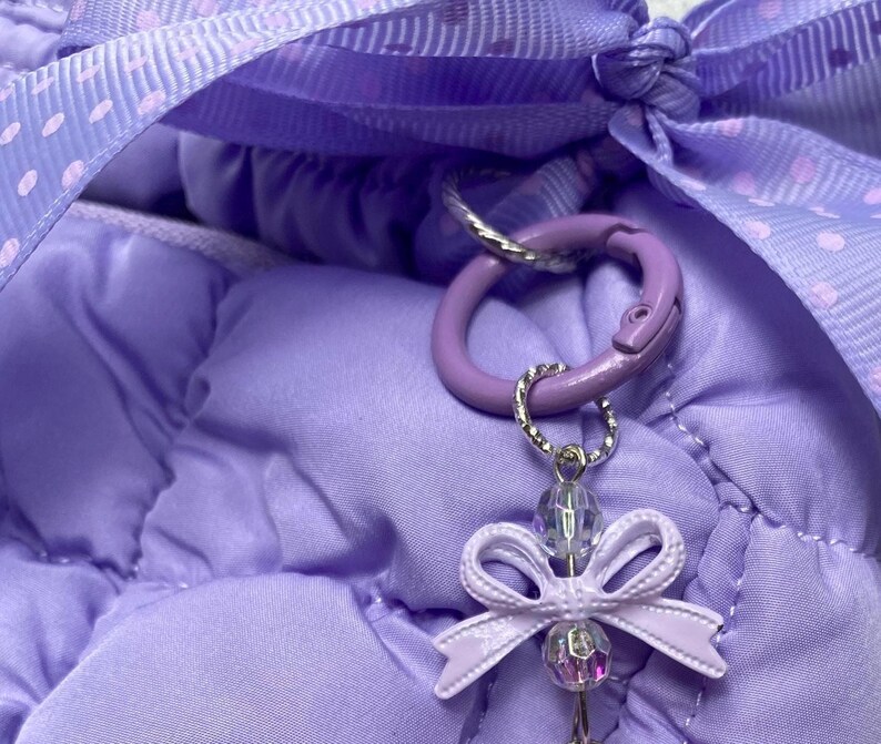 May include: Close-up of a lavender-colored bag with a matching ribbon and a decorative charm. The charm features a small bow and iridescent beads, attached to a purple carabiner clip. The bag has a quilted texture.