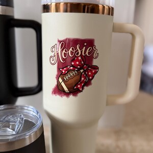 May include: Cream-colored travel mug with a handle, a white lid, and a gold rim. The mug features a graphic of the state of Indiana with the word "Hoosier" and a football with a red polka-dot bow. Other mugs are visible in the background.