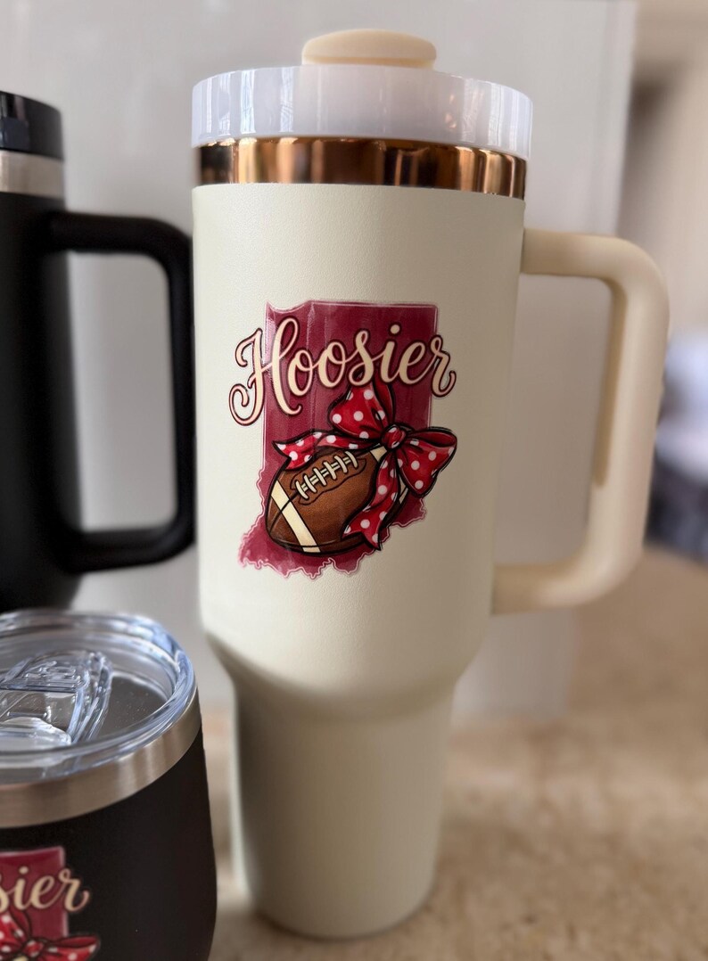 May include: Cream-colored travel mug with a handle, a white lid, and a gold rim. The mug features a graphic of the state of Indiana with the word "Hoosier" and a football with a red polka-dot bow. Other mugs are visible in the background.